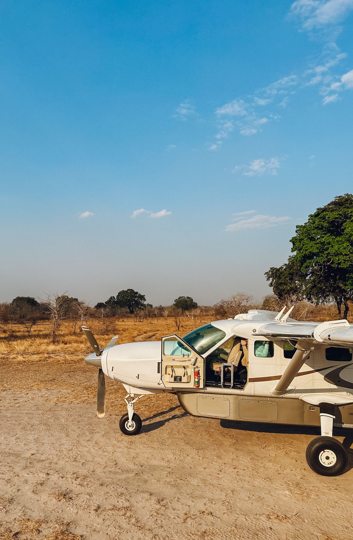 A single-engine bush aircraft parked on a dry dirt savanna airstrip fringed with acacia scrub under a clear sky in Africa